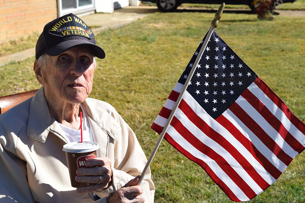 An elderly man with a WWII cap, an American flag and a cup of coffee sitting in his front lawn